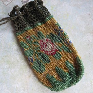 May include: A small drawstring pouch crafted from tiny glass beads in shades of gold, green, and red, featuring a floral pattern. The top section is made of dark olive green crocheted thread with a matching drawstring cord for closure.