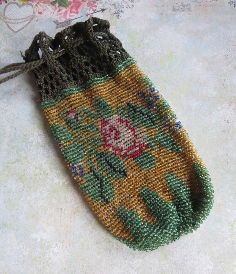 May include: A small drawstring pouch crafted from tiny glass beads in shades of gold, green, and red, featuring a floral pattern. The top section is made of dark olive green crocheted thread with a matching drawstring cord for closure.