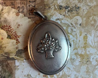 May include: An oval-shaped silver-toned metal locket featuring a raised relief design of a basket filled with flowers on the front surface. The locket includes a small metal bail at the top for attachment to a chain or cord, with a smooth finish.