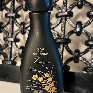 May include: Black bottle of Shiseido Zen Eau de Cologne. The bottle has a rounded top and a black ball-shaped stopper. Gold floral design and text are printed on the front. The background features a decorative black metalwork.