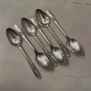 May include: Six silver spoons arranged diagonally. Each spoon has a floral design on the handle and a reflective, oval bowl. The spoons are set against a light gray background, suggesting a dining or serving context.