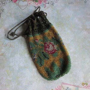May include: A small drawstring pouch crafted from tiny glass beads in shades of gold, green, and red, featuring a floral pattern. The top section is made of dark olive green crocheted thread with a matching drawstring cord for closure.
