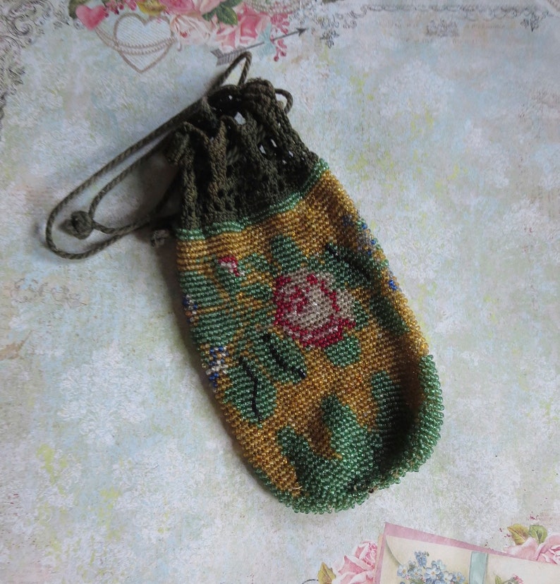 May include: A small drawstring pouch crafted from tiny glass beads in shades of gold, green, and red, featuring a floral pattern. The top section is made of dark olive green crocheted thread with a matching drawstring cord for closure.