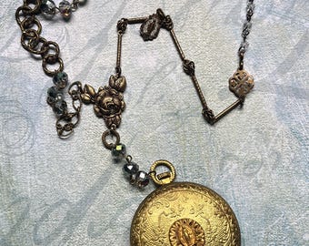 May include: A round, gold-toned metal pocket watch with an ornate floral pattern and a central embossed figure, attached to a long chain featuring dark metallic rings, clear faceted beads, a rose-shaped metal link, and a small religious charm.