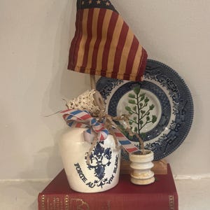 May include: A white ceramic jug with blue text reading FORTE AU VIN BLANC, decorated with a red, white, and blue striped ribbon and a small American flag. It sits on a red book next to a small white stand with a green plant and a blue patterned plate.