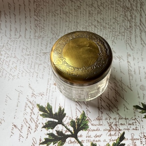 May include: A clear glass jar with a gold-colored lid. The lid has a decorative leaf pattern around the edge and a smooth, circular center. The jar sits on a background of vintage script and botanical illustrations.