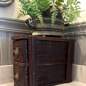 May include: A dark brown wooden storage unit with two stacked drawers, each featuring a textured grid pattern and a circular metal ring pull handle. A metallic, perforated planter containing a green fern sits on top of the wooden unit.