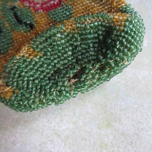 May include: A small pouch crafted from tiny glass beads in shades of emerald green, amber, and red. The surface features a dense, textured pattern of these beads stitched together, creating a rounded shape with a visible opening at the top edge.