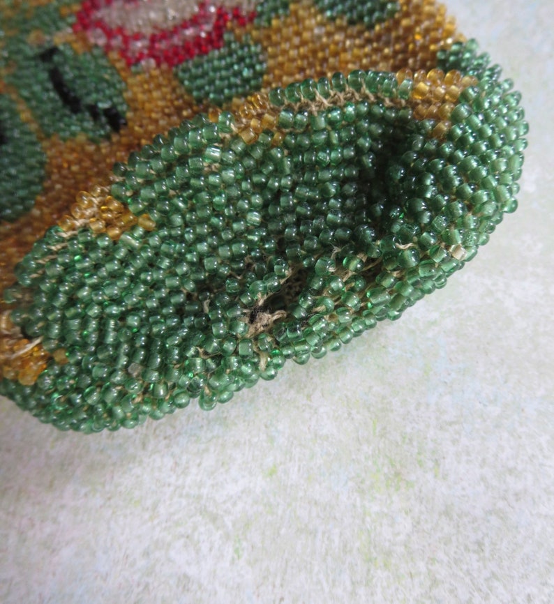 May include: A small pouch crafted from tiny glass beads in shades of emerald green, amber, and red. The surface features a dense, textured pattern of these beads stitched together, creating a rounded shape with a visible opening at the top edge.