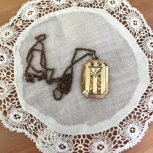 May include: A rectangular gold-tone metal locket pendant with a hinged design, suspended from a matching thin metal chain. The front of the locket features an engraved triangular motif containing the letter F, flanked by vertical decorative patterns.