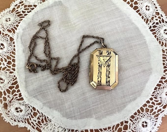 May include: A rectangular gold-tone metal locket pendant with a hinged design, suspended from a matching thin metal chain. The front of the locket features an engraved triangular motif containing the letter F, flanked by vertical decorative patterns.