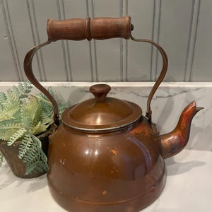 May include: A copper-colored tea kettle with a wooden handle and lid knob. The kettle has a curved spout and a metal handle that arches over the top. The kettle is sitting on a white surface.
