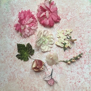 May include: A collection of decorative floral elements including two large pink fabric flowers, a pale cream blossom, a cluster of small white flowers, a green textured leaf, a dried rosebud, a small beige flower, a sprig of greenery, and beaded pink buds.