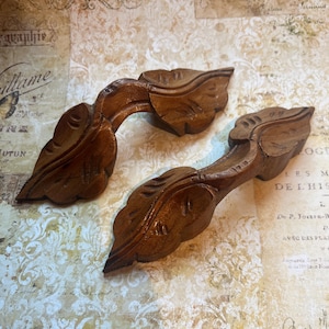 May include: Two carved wooden leaf-shaped decorative elements. The pieces are a warm brown color and feature detailed veining and a curved design. They appear to be vintage or antique, possibly for furniture or decor.