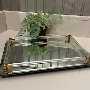 May include: A rectangular mirrored tray featuring a reflective glass base and a clear glass railing supported by four metallic gold-toned corner connectors. The tray has a clean, polished appearance with a smooth surface and structured edges.