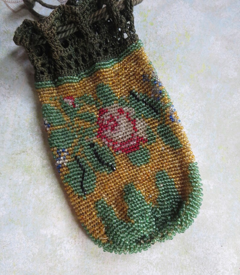 May include: A drawstring pouch featuring a dark olive green crocheted top and a body covered in intricate glass beads. The beadwork displays a floral pattern with a central pink rose, green leaves, and a golden yellow background with green trim.