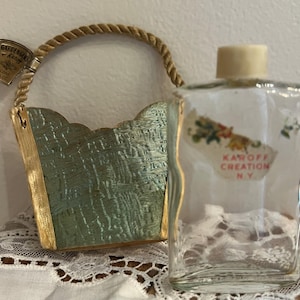 May include: A vintage perfume bottle with a clear glass body and a white plastic cap. The bottle has a label that reads "Kamoff Creation N.Y.". The bottle is sitting next to a small, green and gold box with a rope handle.