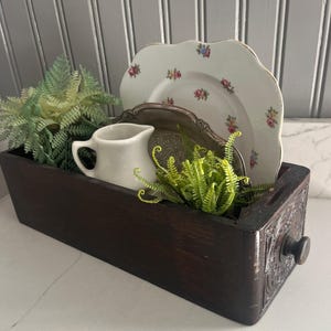 May include: A dark brown wooden drawer box containing a white ceramic creamer, a silver-toned metal tray, a white plate with a scalloped edge and floral pattern, and two green artificial fern plants placed inside the rectangular container.