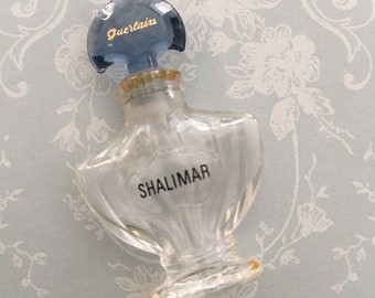 May include: A clear glass bottle of Shalimar perfume by Guerlain. The bottle has a blue and gold cap and a textured design. The bottle is sitting on a light blue background with a floral pattern.