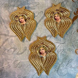 May include: Three gold embroidered angel appliques. Each applique features a portrait of a cherubic face with a floral crown, surrounded by gold wings. The appliques are embellished with small, sparkling rhinestones.