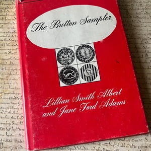 May include: A red book titled "The Button Sampler" with white text and illustrations of buttons. The book cover also features the authors' names, Lillian Smith Albert and Jane Ford Adams, in elegant script.