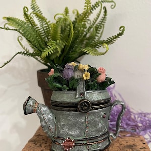 May include: A decorative, miniature watering can-shaped container with a hinged lid, filled with colorful artificial flowers. The container is painted in shades of gray and has a handle and spout. A potted fern is in the background.