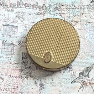 May include: A vintage, round, gold-toned compact with a ribbed design on the lid. The compact features a small, oval emblem near the bottom. The background is a collage of vintage paper with script and illustrations.