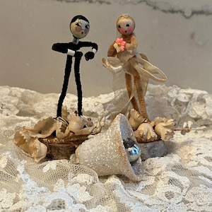 May include: A vintage wedding cake topper featuring two handmade figures. The groom is in a black suit, and the bride wears a veil and holds a pink flower. A bell-shaped ornament with a silver ball is also present.