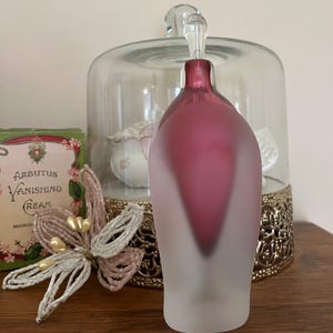 May include: A frosted glass bottle with a pink and white gradient. The bottle has a unique shape with a pointed bottom and a rounded top. The bottle is sitting on a gold decorative stand.