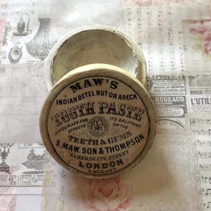May include: A round, off-white ceramic pot with a removable lid. The lid features black printed text reading: MAW'S INDIAN BETEL NUT OR ARECA TOOTH PASTE, UNEQUALLED FOR EFFECTS, ITS SALUTARY ON THE TEETH & GUMS, S. MAW, SON & THOMPSON, LONDON, ENGLAND.