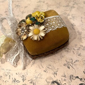 May include: Mustard-colored velvet pincushion with white lace trim and floral embellishments. The pincushion is adorned with a cluster of vintage-style flowers in shades of yellow, cream, and gold. A white lace ribbon is attached for hanging.