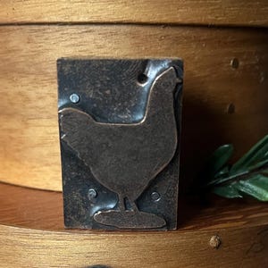 May include: A rectangular metal printing block featuring a raised silhouette of a chicken in profile. The dark, weathered metal surface shows a textured finish with three small circular rivets visible on the face of the block surrounding the bird.