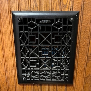 May include: A rectangular black metal vent cover with a geometric pattern of intersecting lines and shapes. The vent is mounted on a wooden surface with vertical grain. The vent is secured with four screws.