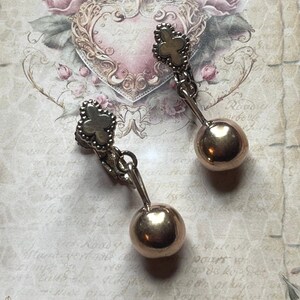 May include: A pair of gold tone clip-on earrings with a dangling ball charm. The earrings feature a clover-shaped design with a textured surface.