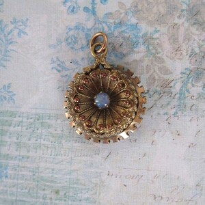 May include: A gold-toned pendant with a central blue gemstone and intricate floral detailing. The pendant features a circular design with a decorative outer edge and small red accents. It has a loop at the top for hanging.