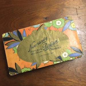 May include: Vintage box of Normandy Puritan Chocolates, featuring a gold oval label with the brand name and the text "Fruits, Nuts and Creams" and "One Pound Net". The box has a colorful art deco design with orange, green, blue, and gold accents.