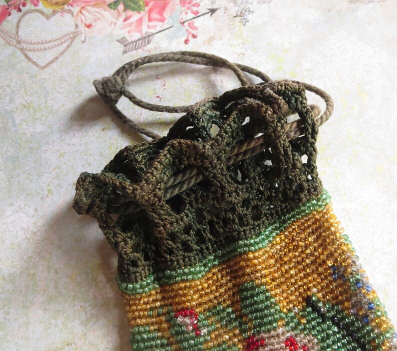 May include: A small drawstring pouch featuring a dark olive green crocheted top section with a matching cord closure. The lower portion of the pouch is covered in intricate, multi-colored glass seed beads in shades of gold, green, and red.