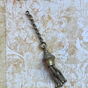 May include: A vintage-style tassel pendant with a decorative cap and braided strands. The pendant is attached to a chain and ring, and is made of a gold-toned metal. The background is a patterned, off-white surface.
