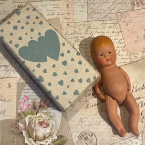 May include: A vintage doll with a painted face and limbs, next to a rectangular box. The box is beige with a pattern of small blue hearts and two larger hearts. The doll is lying on a background of vintage postcards.