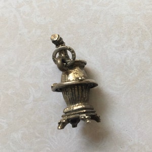 May include: A small, silver-toned charm in the shape of a vintage samovar. The charm features a detailed design with a cylindrical body, a flared base, and a small spout. A ring is attached to the top for hanging. The charm is likely made of metal.