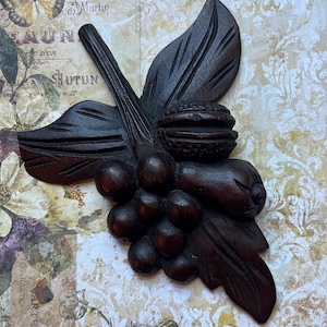 May include: A dark brown, carved wooden decorative piece featuring a cluster of grapes, leaves, and a nut. The intricate carving showcases detailed textures and shapes, creating a rustic, natural aesthetic. The piece is likely a wall hanging or decorative element.