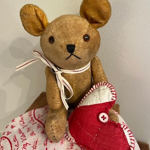 May include: A brown teddy bear with large ears and brown button eyes is holding a red heart-shaped pillow with white stitching and a red button. The pillow is sitting on a white fabric with red writing that says "Be My Valentine".