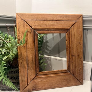 May include: A rectangular picture frame constructed from medium-toned brown wood with a visible grain pattern. The frame features mitered corners and a stepped, beveled profile that slopes inward toward the central open rectangular space.