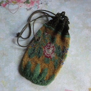 May include: A small drawstring pouch covered in intricate glass beads. The beadwork features a floral pattern with a central pink rose, green leaves, and a mustard yellow background. The top of the pouch has a dark green crocheted closure with matching cords.