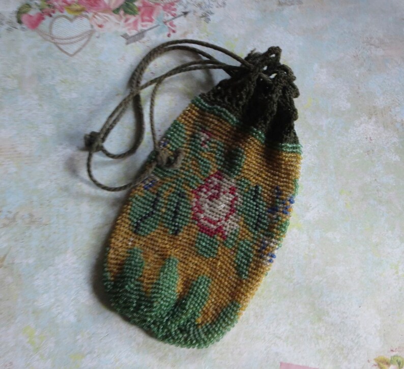 May include: A small drawstring pouch covered in intricate glass beads. The beadwork features a floral pattern with a central pink rose, green leaves, and a mustard yellow background. The top of the pouch has a dark green crocheted closure with matching cords.