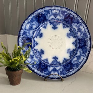 May include: A round ceramic plate featuring a deep blue and white transferware pattern with ornate scrollwork and floral motifs along the scalloped rim, surrounding a central white area with a subtle, light-colored decorative medallion design.
