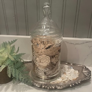 May include: A clear glass apothecary jar filled with vintage lace, doilies, and other craft supplies. The jar sits on a silver tray with a collection of white buttons. A fern in a bronze pot is on the left.