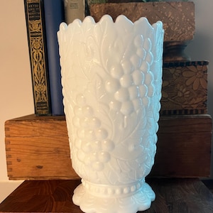 May include: A white, vintage-style vase with a detailed grape and leaf pattern. The vase has a scalloped rim and a pedestal base. The design is embossed on the opaque glass. The vase is approximately 8 inches tall.