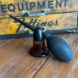 May include: An antique brown glass atomizer with a black rubber bulb and nozzle. The atomizer sits on a wooden surface in front of a vintage wooden cabinet with the word "EQUIPMENT" visible.