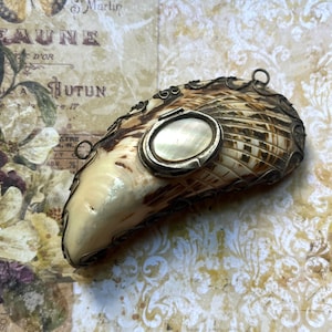 May include: A curved, cream and brown patterned seashell pendant or brooch encased in a decorative, dark metal wire filigree frame. The center features an oval, iridescent mother-of-pearl inlay set within a smooth, polished metal bezel.